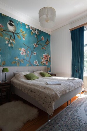 A bedroom with a large floral mural, a bed with green pillows, a white throw, and a chandelier. - Amiralsgården Werstorp (Nättraby)