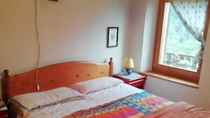 A bedroom with a wooden bed, a colorful quilt, a bedside table with a lamp, and a window with a view of greenery. - Mountain style apartment (Zortea)