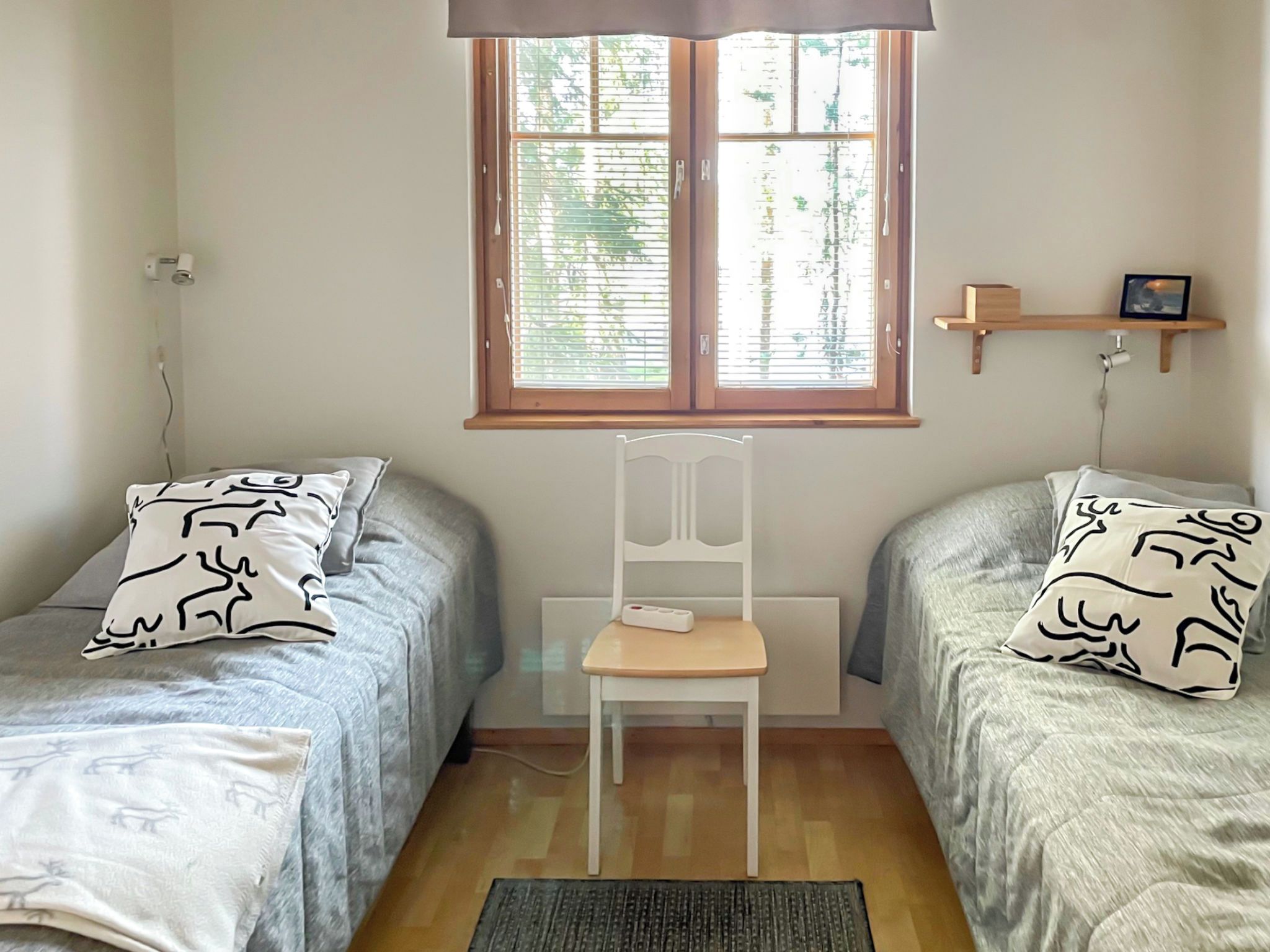 A room with two single beds, a chair, a window with blinds, a shelf with a picture, and a radiator.