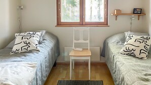 Habitación con dos camas, una silla, una ventana con persianas, un estante con un cuadro y un enchufe.