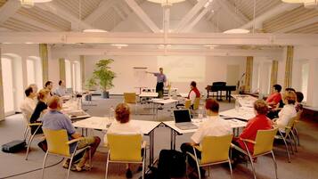 Ein Konferenzraum mit Präsentationseinrichtung, darunter eine Whiteboard, eine Projektionsleinwand und ein Referent. Teilnehmer sitzen an Tischen mit Laptops und Notizbüchern.
