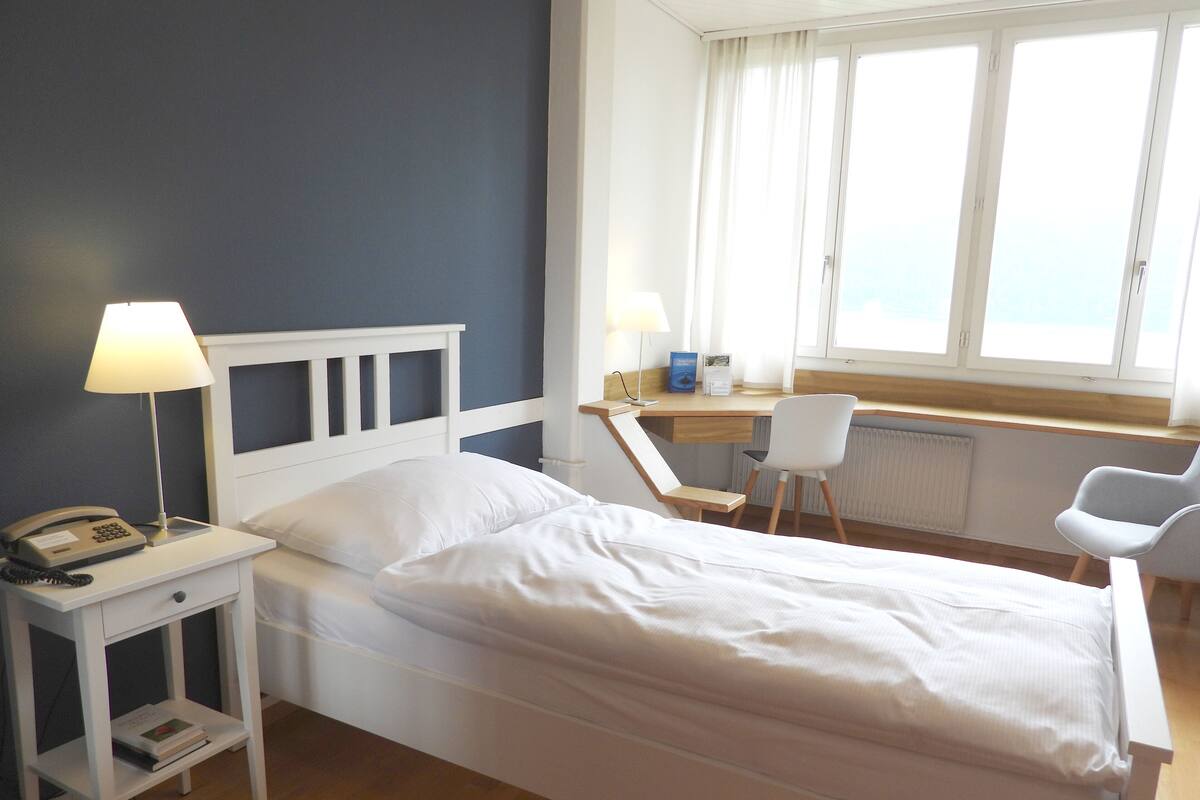 A single bed with a white headboard, a bedside table with a lamp, a chair, and a desk with a chair.