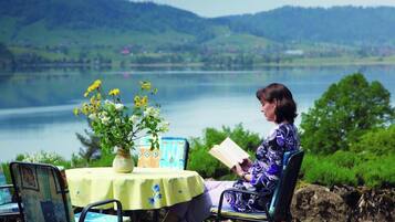 Una persona sentada a una mesa con un libro, contemplando un lago con colinas al fondo.