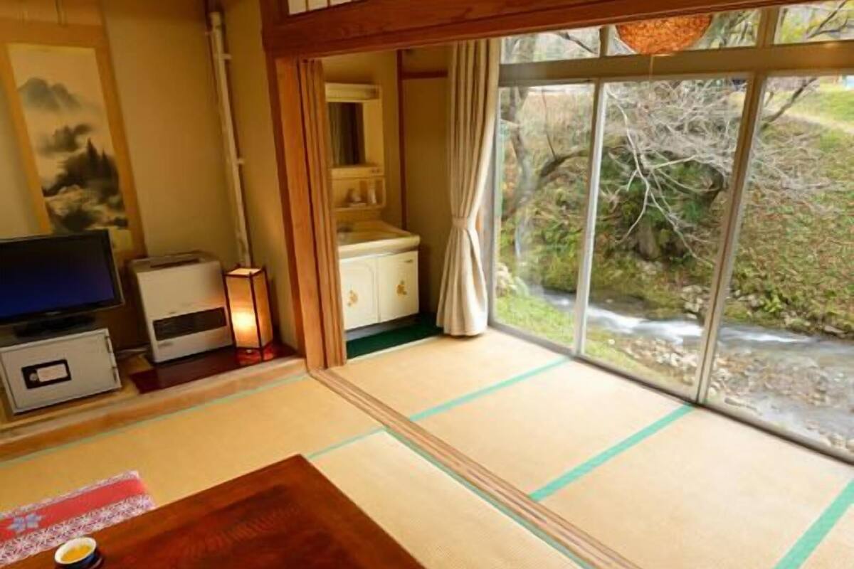 Um espaço com esteiras de tatame, uma mesa de madeira, televisão e vista para um jardim através de uma porta deslizante.