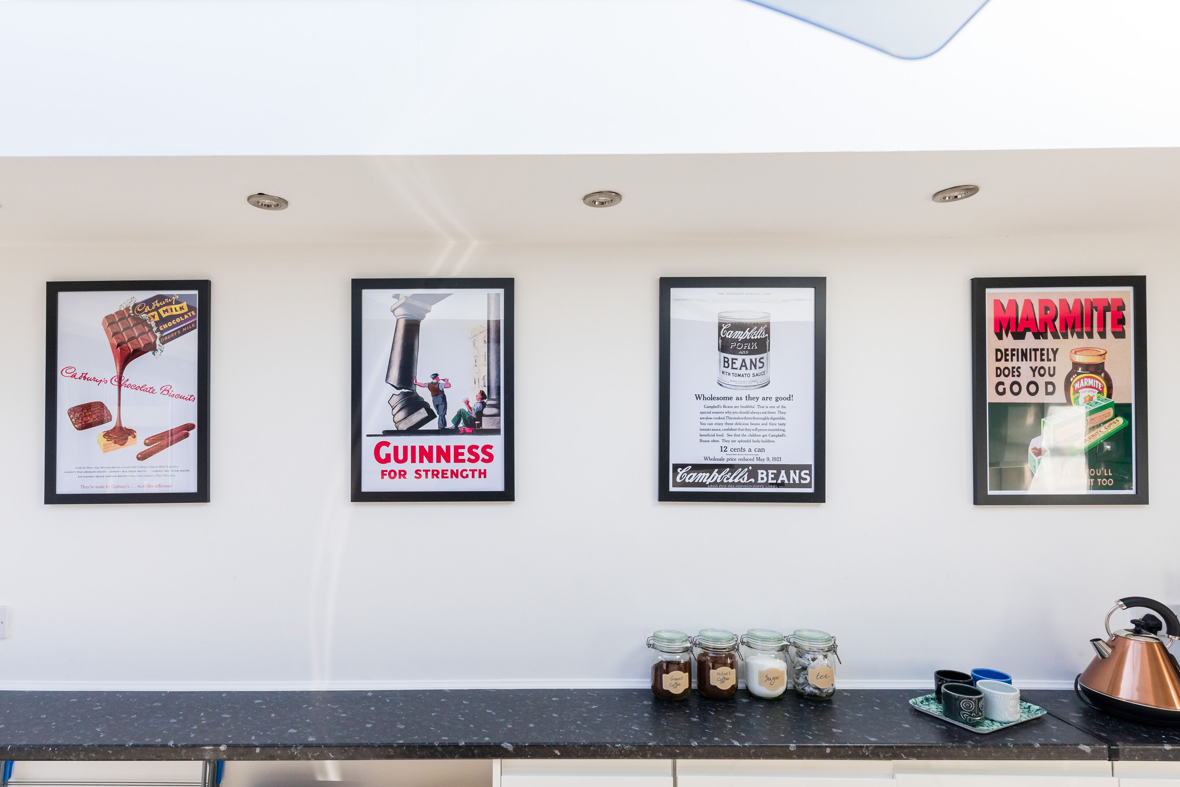 A kitchen wall with four framed advertisements: Guinness for strength, Campbell's beans, Marmite, and a chocolate advertisement. There are jars of jam and a kettle on the counter.