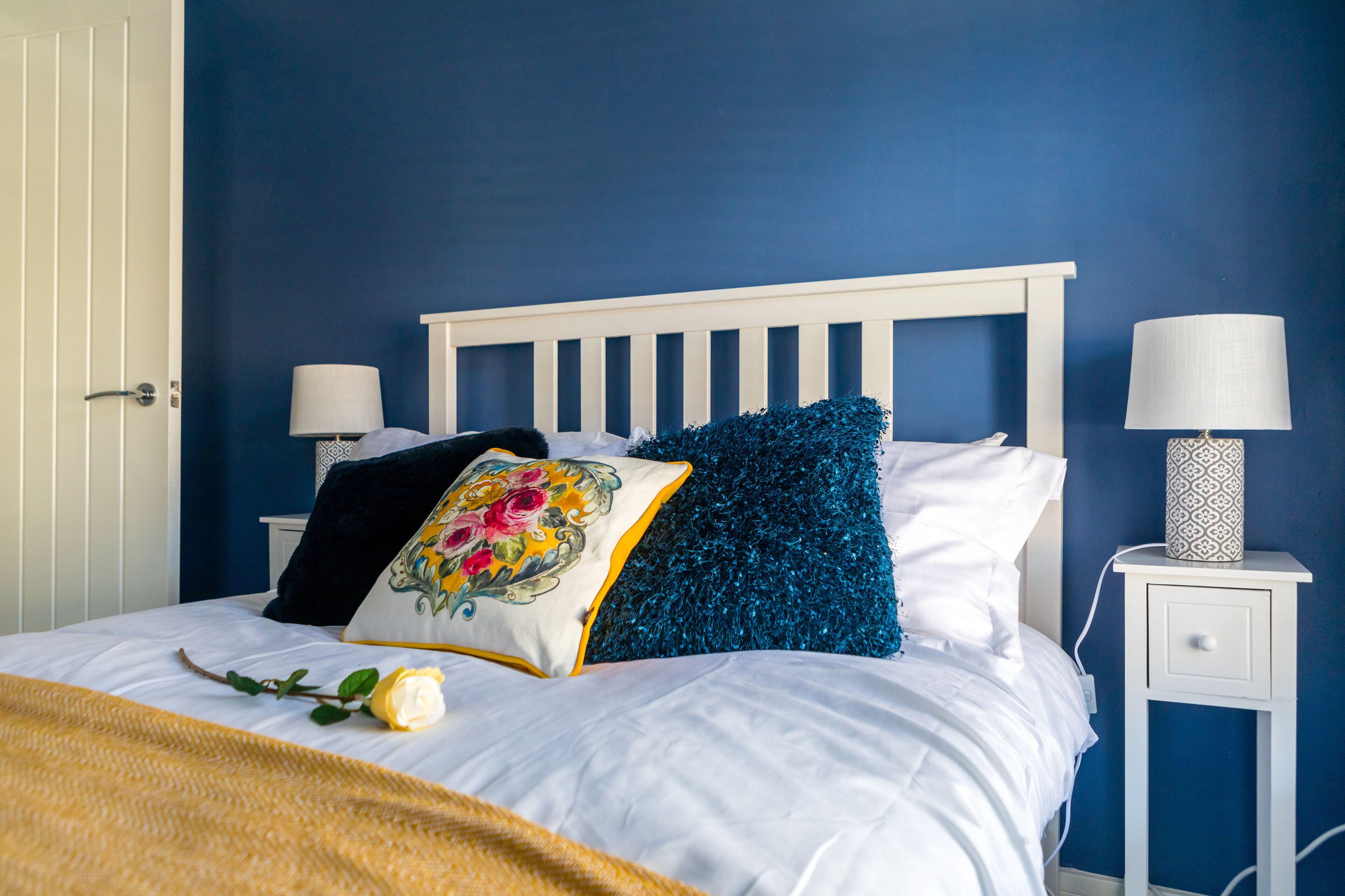 Een netjes opgemaakt bed met witte beddengoed en decoratieve kussens, een nachtkastje met een lamp, en een deur aan de linkerzijde.