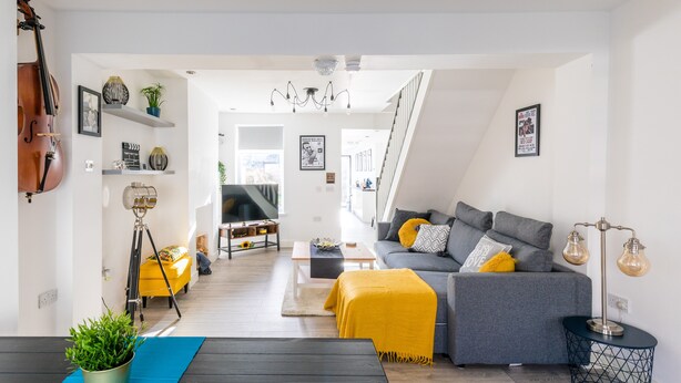 A modern living room with a sofa, coffee table, and a yellow ottoman. There is a cello on the left and a tripod lamp. - The Hollywood Beautiful 3 Bedroom House (Cardiff)