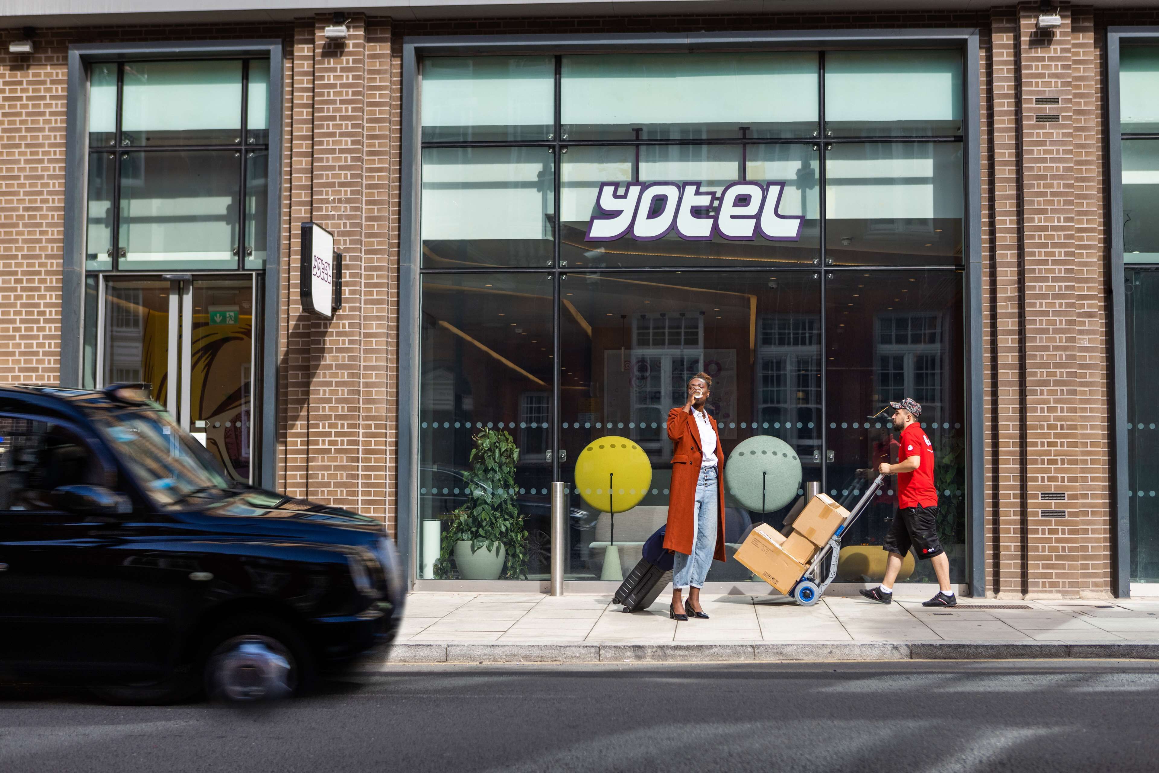 En hotellentrél med en stor glasfasad, en skylt med texten ”yotel”, och en person som skjuter en bagagevagn.