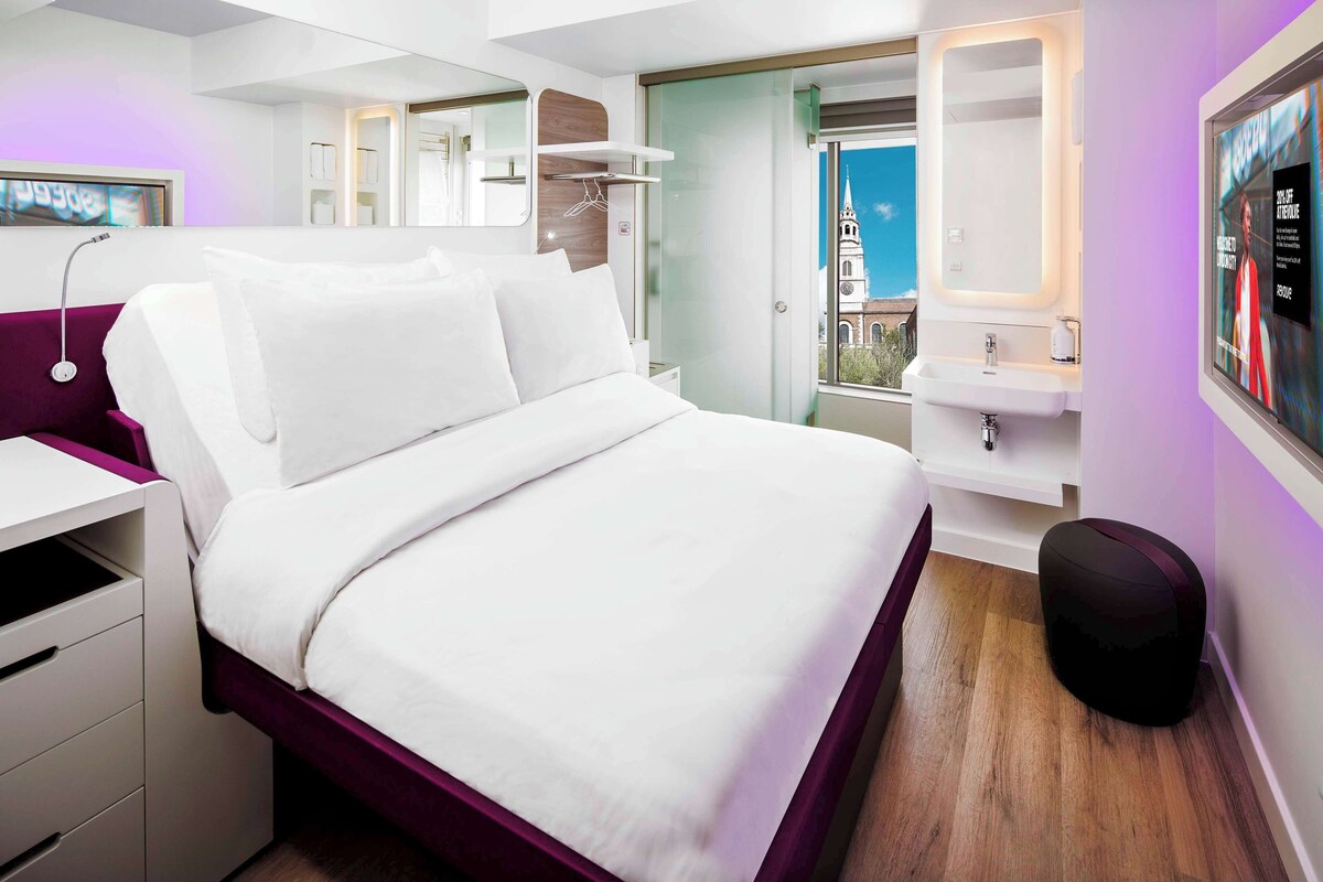 A modern hotel room with a bed, a desk, a vanity with a mirror, and a window with a view of a building.