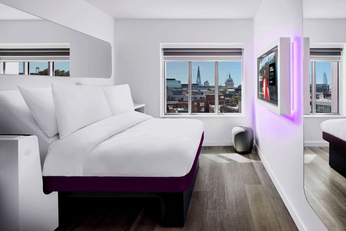 A modern bedroom with a large bed, a TV mounted on the wall, and a city view through the windows.