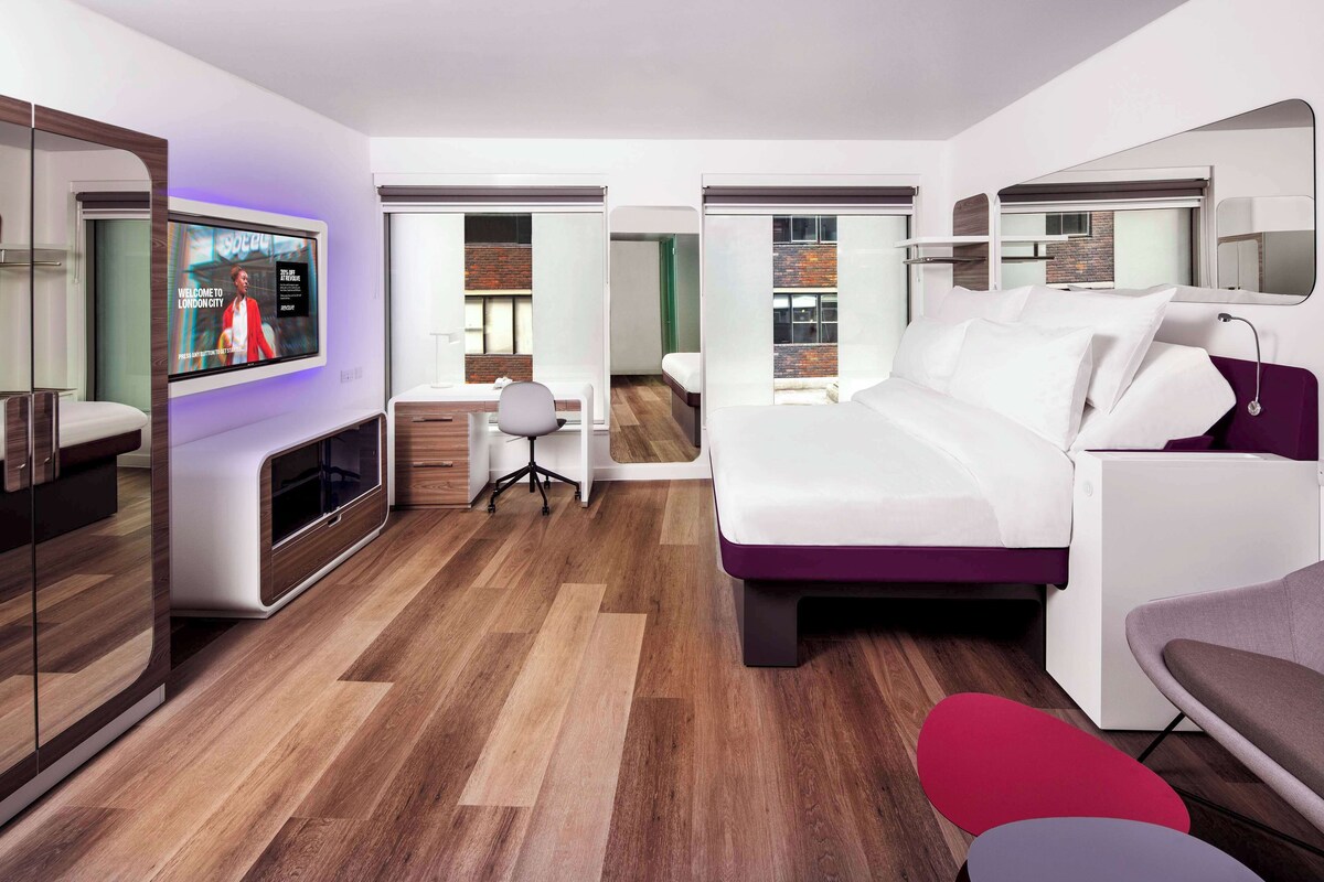 A modern hotel room with a large TV, a bed, a desk, and a chair.