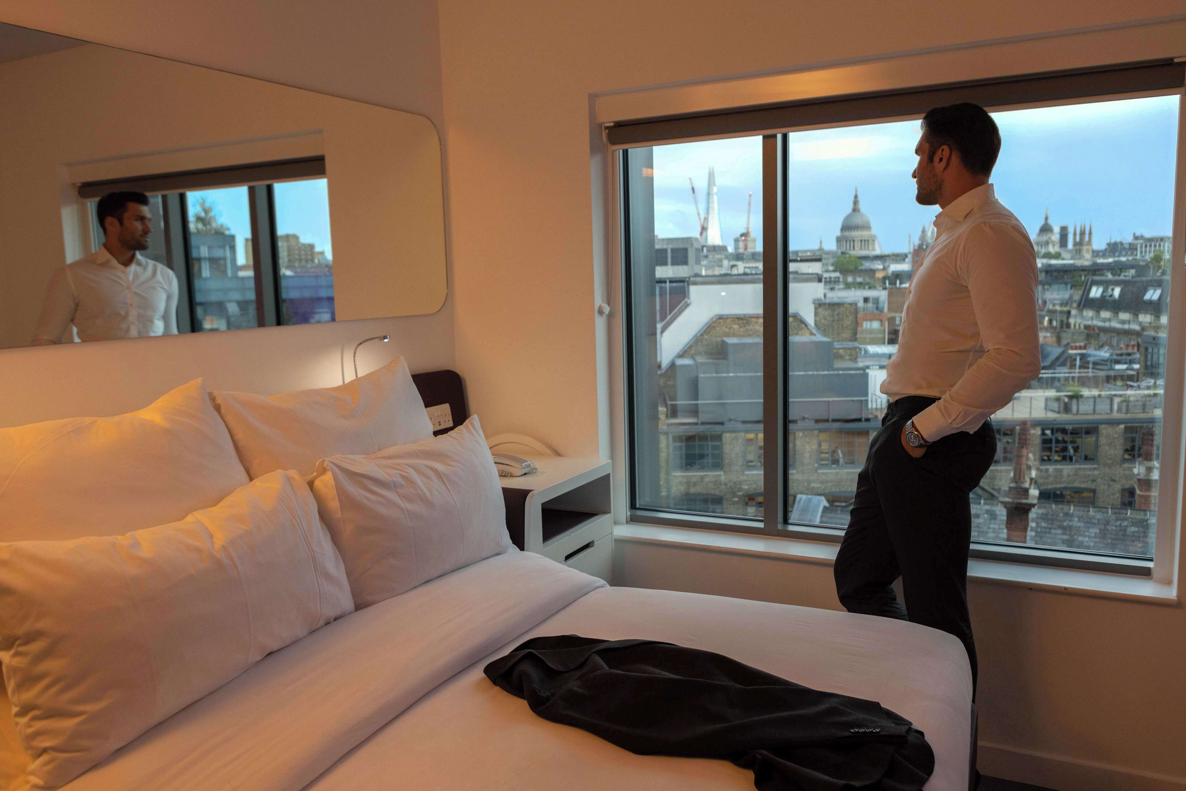 A hotel room with a large bed, a bedside table with a lamp, a mirror, and a window with a city view.