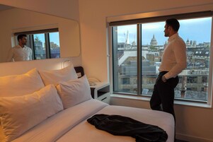 Una habitación de hotel con una cama grande, una mesita de noche con lámpara, un espejo y una ventana con vistas a la ciudad.