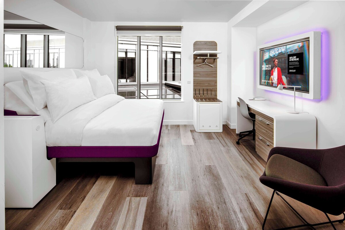 A modern hotel room with a bed, a desk, a chair, a TV, and a wardrobe.