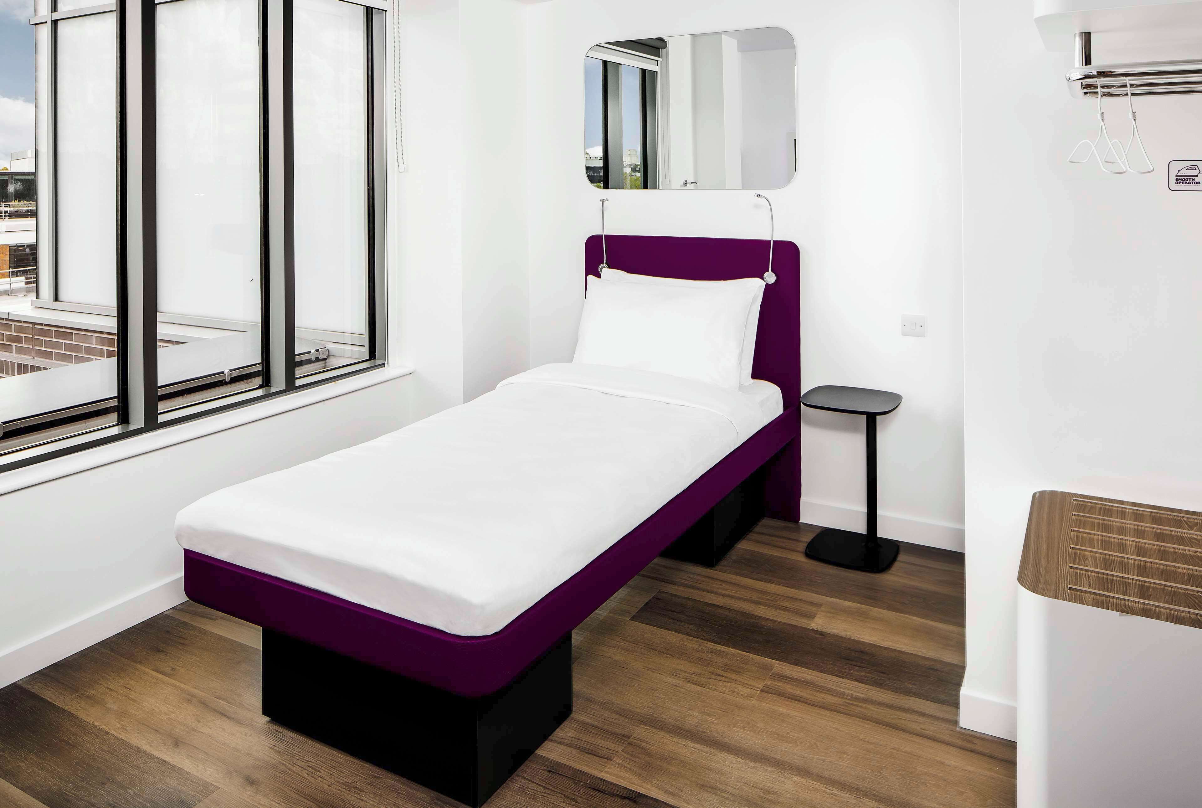 A single-bed room with a purple headboard, a small bedside table, a mirror, and a window with a view of the city.
