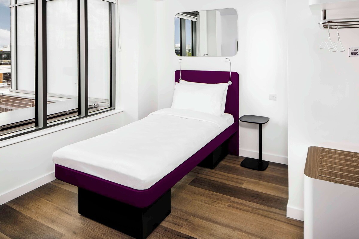 A single-bed room with a purple headboard, a small bedside table, a mirror, and a window with a view of the city.