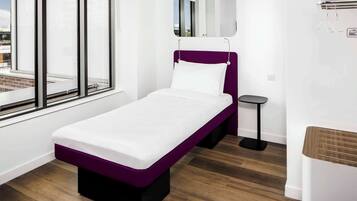 A single-bed room with a purple headboard, a small bedside table, a mirror, and a window with a view of the city.