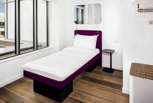 Einzimmerappartement mit einem violetten Kopfteil, einem kleinen Nachttisch, einem Spiegel und einem Fenster mit Blick auf die Stadt.
