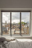 A bedroom with a large window overlooking a cityscape, including historic buildings and a modern skyscraper.