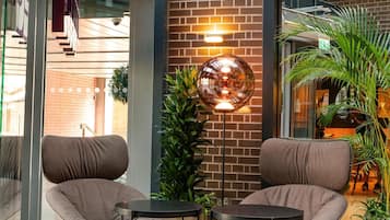 A modern indoor space with brick walls, two cushioned chairs, a small table, a floor lamp, and potted plants.