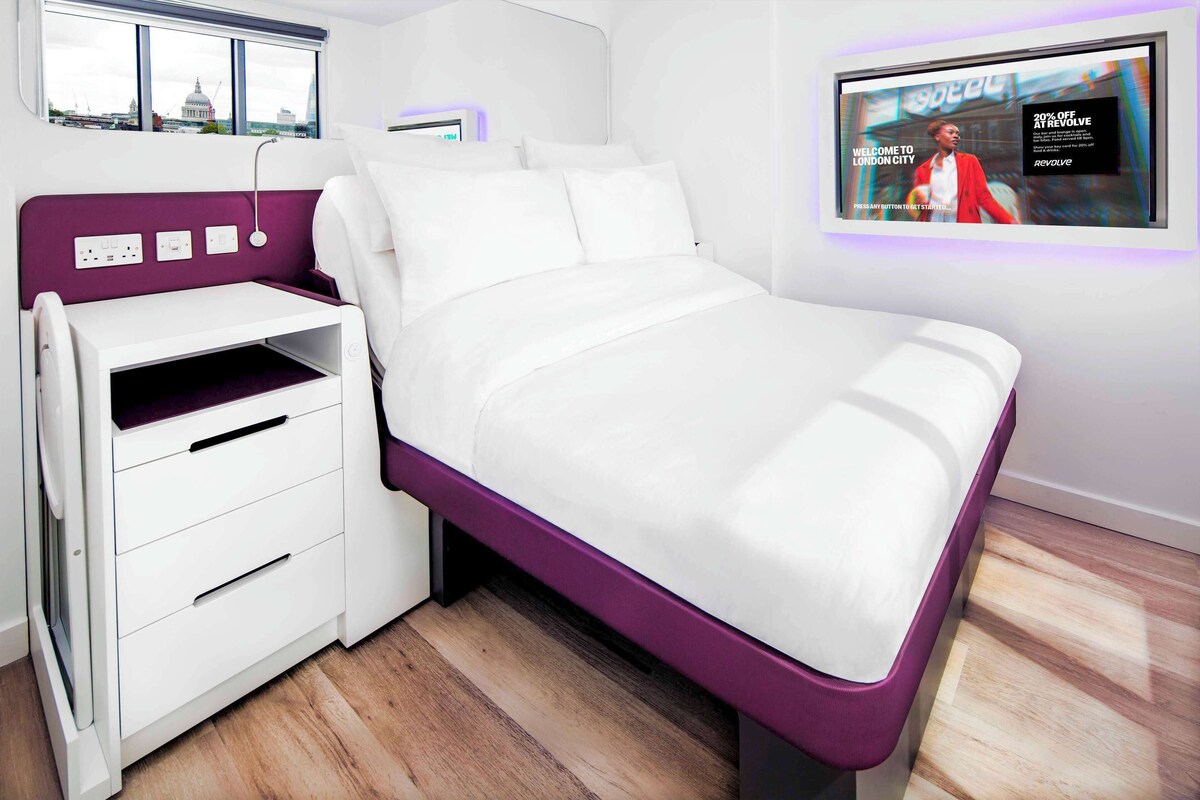 A modern bed with a purple frame and white bedding, a white bedside table with drawers, and a wall-mounted TV displaying a London cityscape.