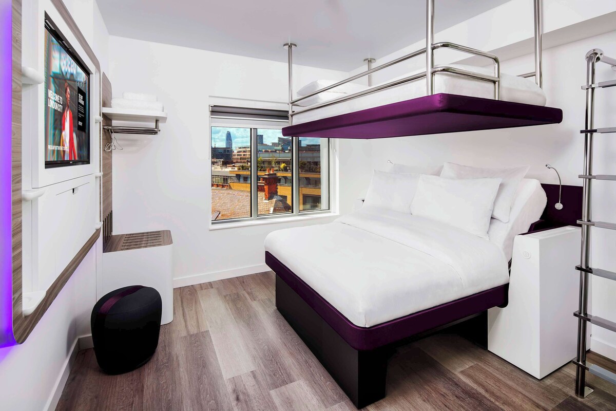 A modern bedroom with a bunk bed, a small desk, and a window with a city view.