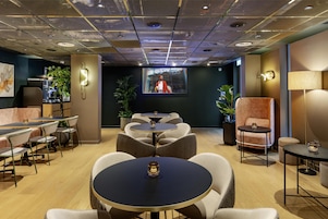 Ein moderner Lounge-Bereich mit runden Tischen, bequemen Sitzgelegenheiten und einem großen Bildschirm, der eine Person zeigt.