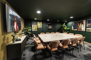 A conference room with a large table, chairs, a plant, and a screen displaying a person.