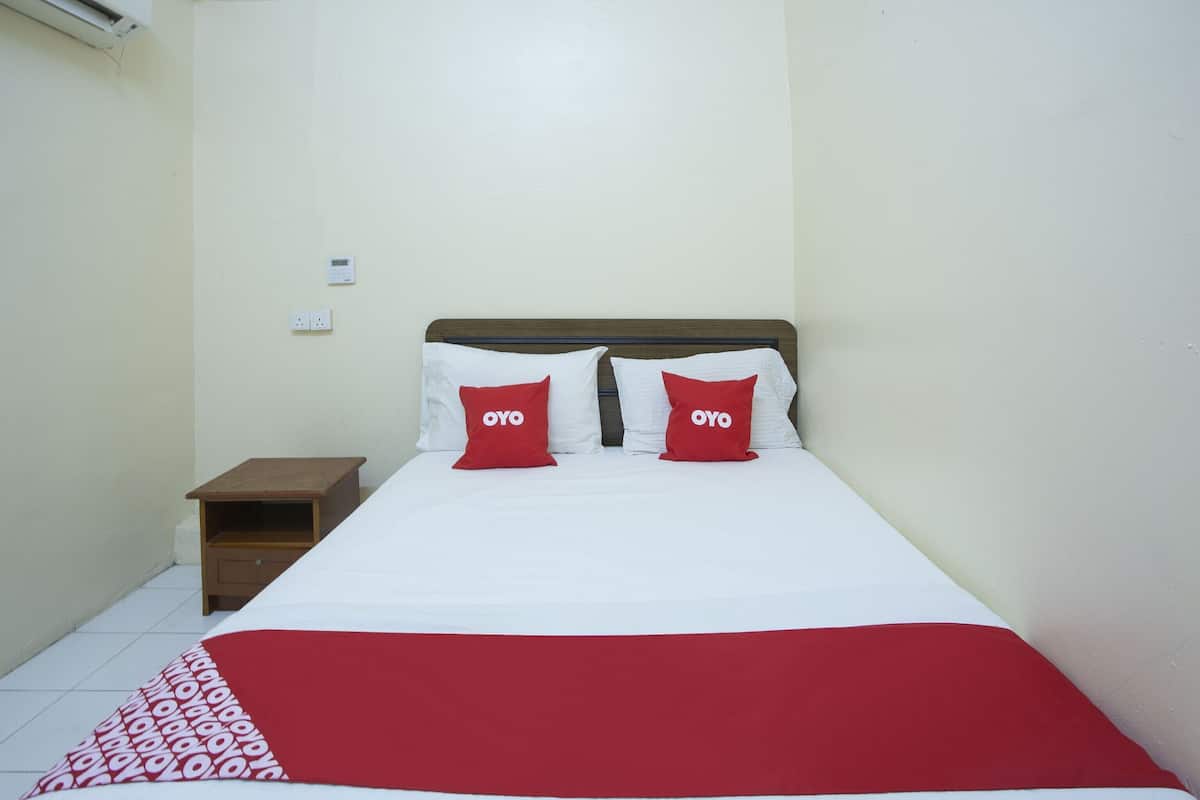 A single bed with white and red bedding, two red pillows with "OYO" text, a wooden nightstand, and a white wall with a power outlet.