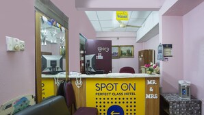 Un espace d’accueil avec une enseigne indiquant « Spot On Perfect Class Hotel », un comptoir de réception, un téléphone, un coin salon avec un fauteuil à motifs verts et une petite table avec un distributeur d’eau.