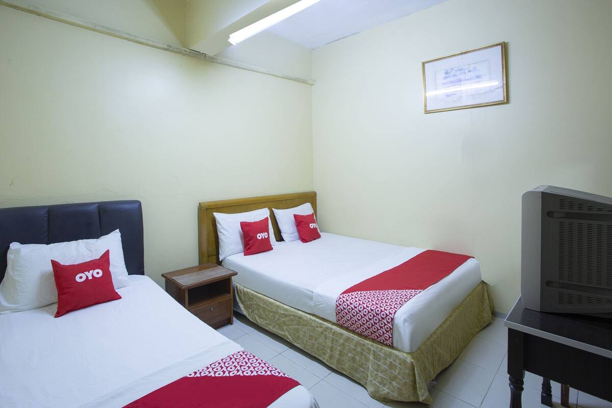 A hotel room with two beds, each with white bedding and red pillows, a wooden headboard, a nightstand, a television, and a framed certificate on the wall.