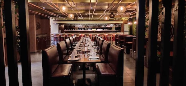 A long dining table with multiple chairs, a bar area in the background, and a modern interior design.