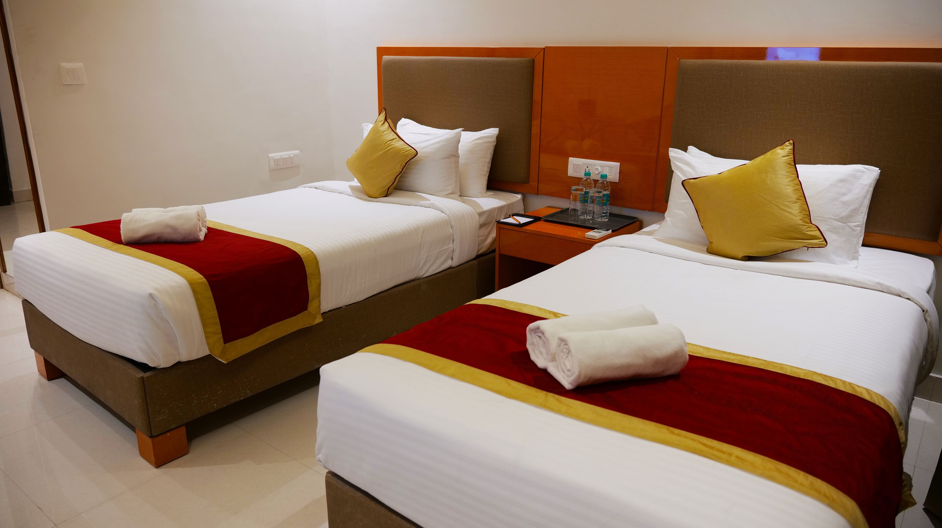 Two neatly made beds with red and white bedding, a nightstand with a lamp, and a wall socket.