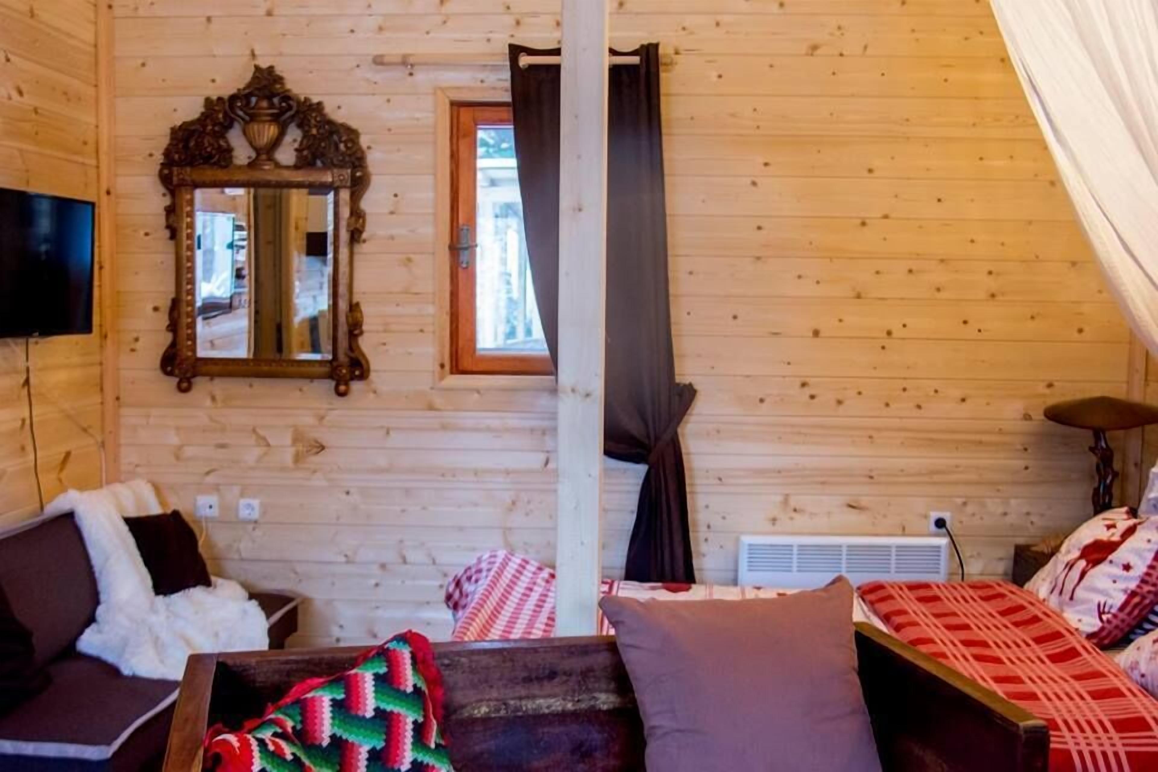 A cozy cabin interior with a wooden ceiling, a large mirror, a sofa, a bed with patterned bedding, and a television on the wall.