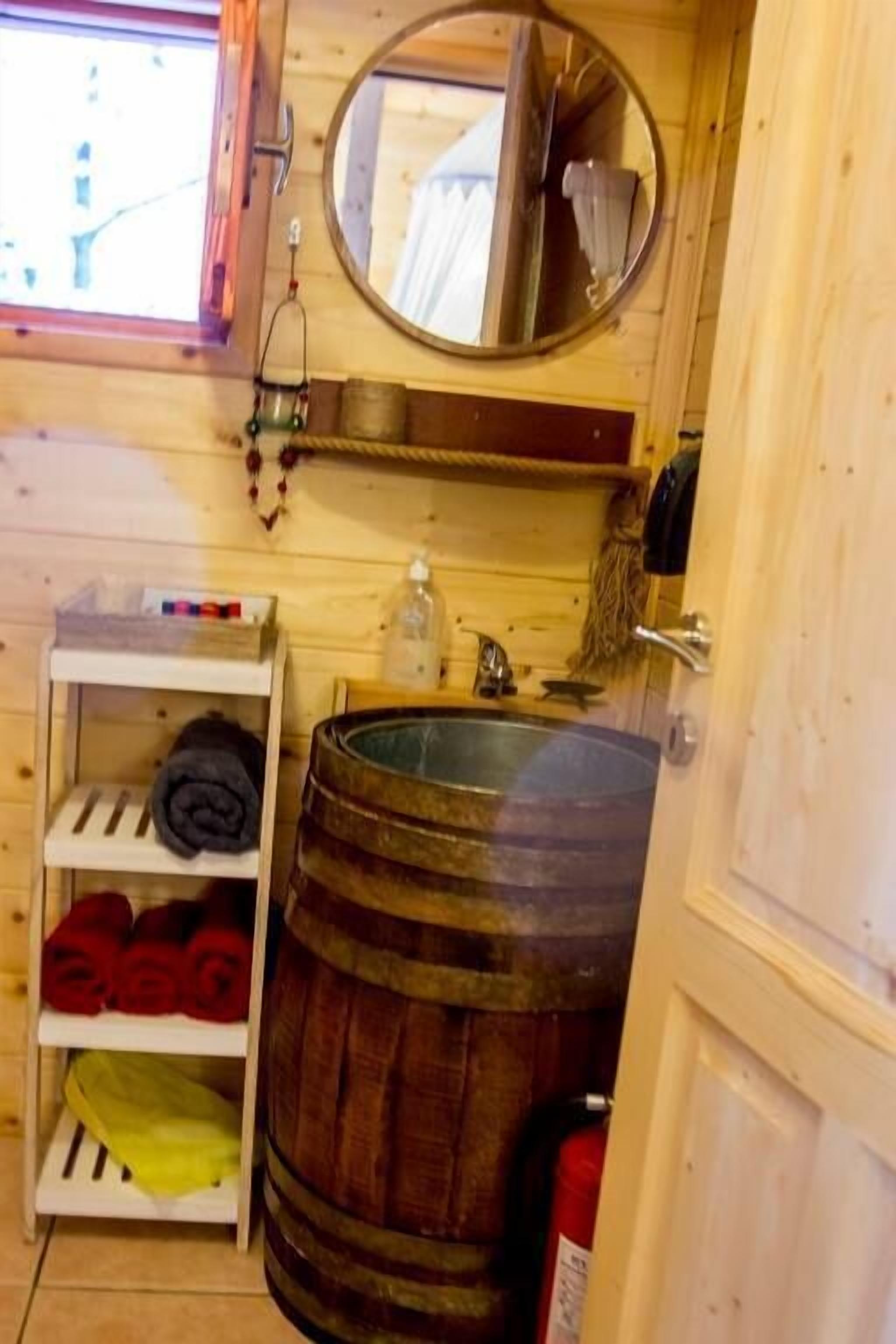 A wooden cabin interior with a wooden shelf holding towels, a round mirror, a wooden barrel, and a sink.