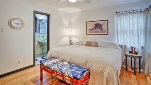 Una camera da letto con un letto, un comodino con libri, una lampada, una ventola a soffitto e una finestra con tende.