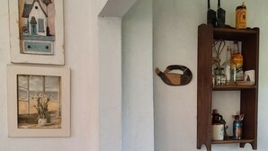 A dining area with a wooden table, a fruit bowl, a wall-mounted shelf with bottles, and two framed pictures.