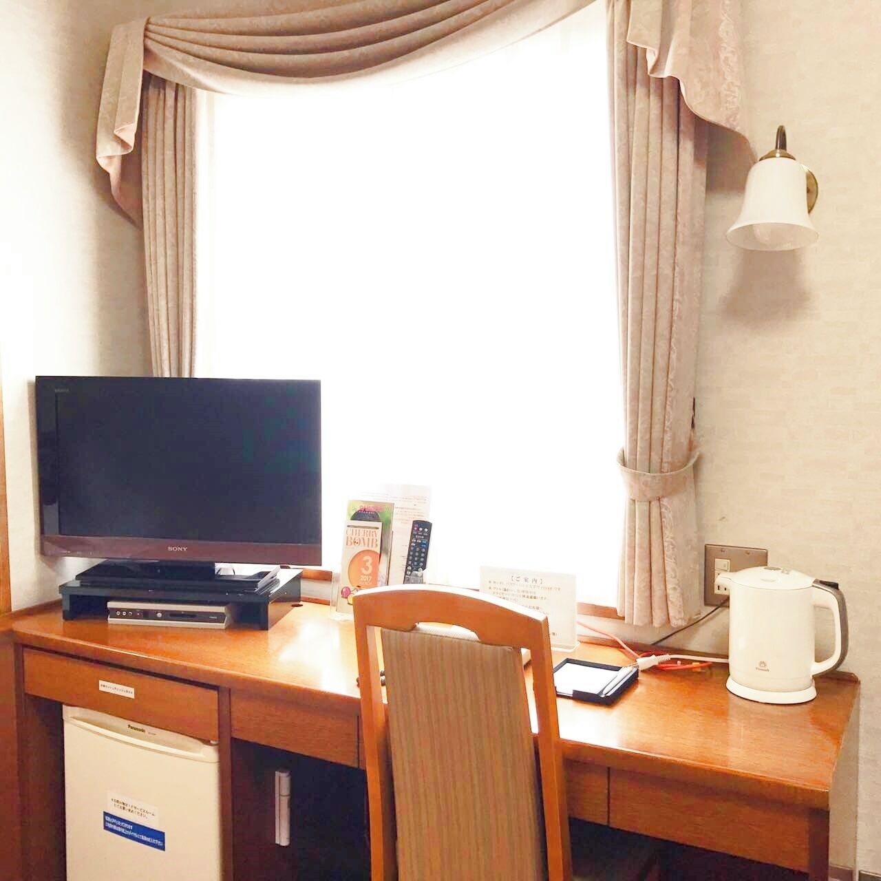 A hotel room with a wooden desk, a television, a coffee maker, a lamp, and a chair.