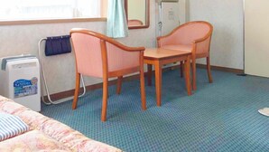 A hotel room with a bed, two wooden chairs, a small table, a window with light blue curtains, a mirror, and a radiator. - Hotel River Inn (Nagaoka)