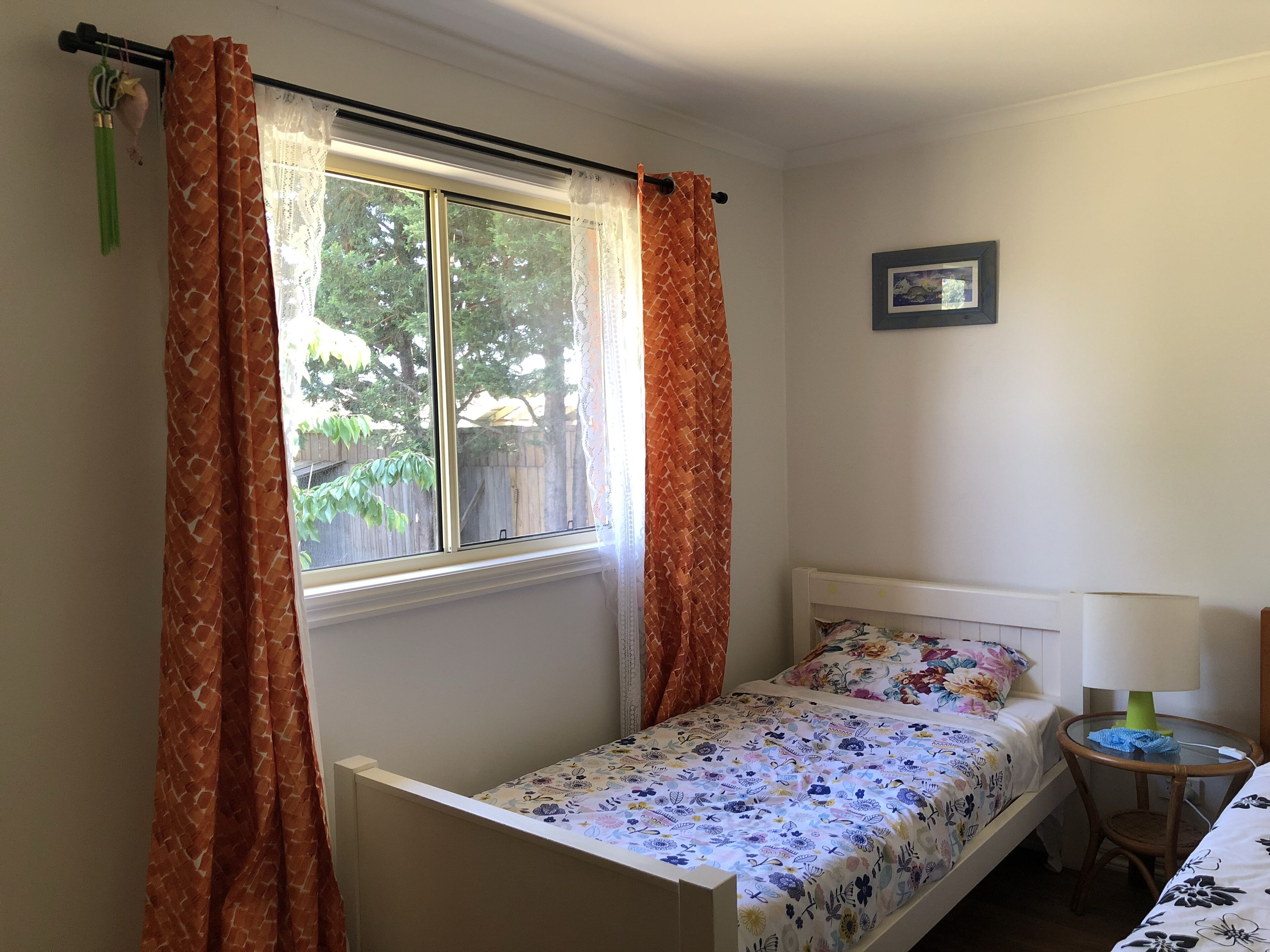 Een slaapkamer met een enkel bed, een klein nachtkastje, een lamp en een raam met gordijnen.