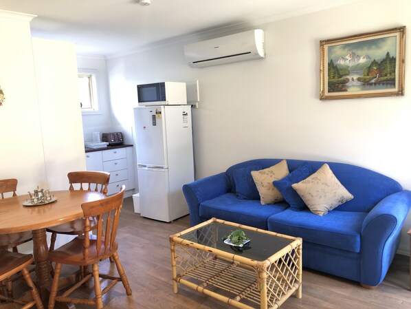 Un espace de vie compact comprenant une table à manger, des chaises, un canapé bleu avec des coussins, un petit coin cuisine équipé d’un micro-ondes et d’un réfrigérateur, et un tableau accroché au mur.