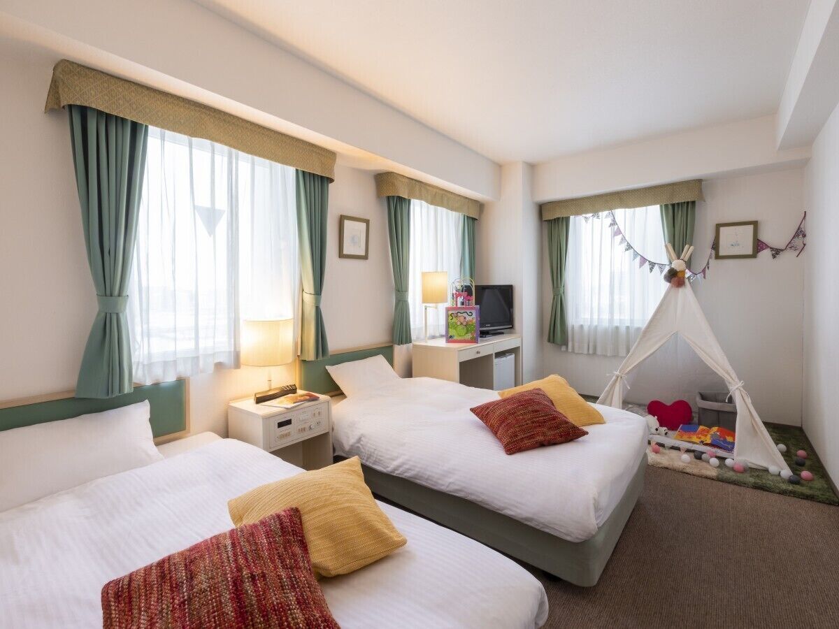 Une chambre d’hôtel avec deux lits, une télévision, une table de chevet, une lampe, un petit espace de jeux avec une tente et un tableau accroché au mur.