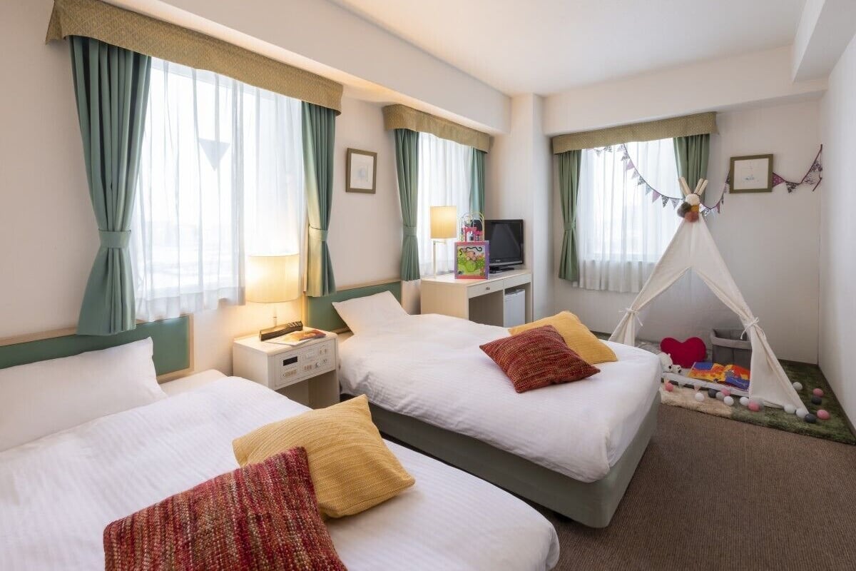 Une chambre d’hôtel avec deux lits, une télévision, une table de chevet, une lampe, un petit espace de jeux avec une tente et un tableau accroché au mur.