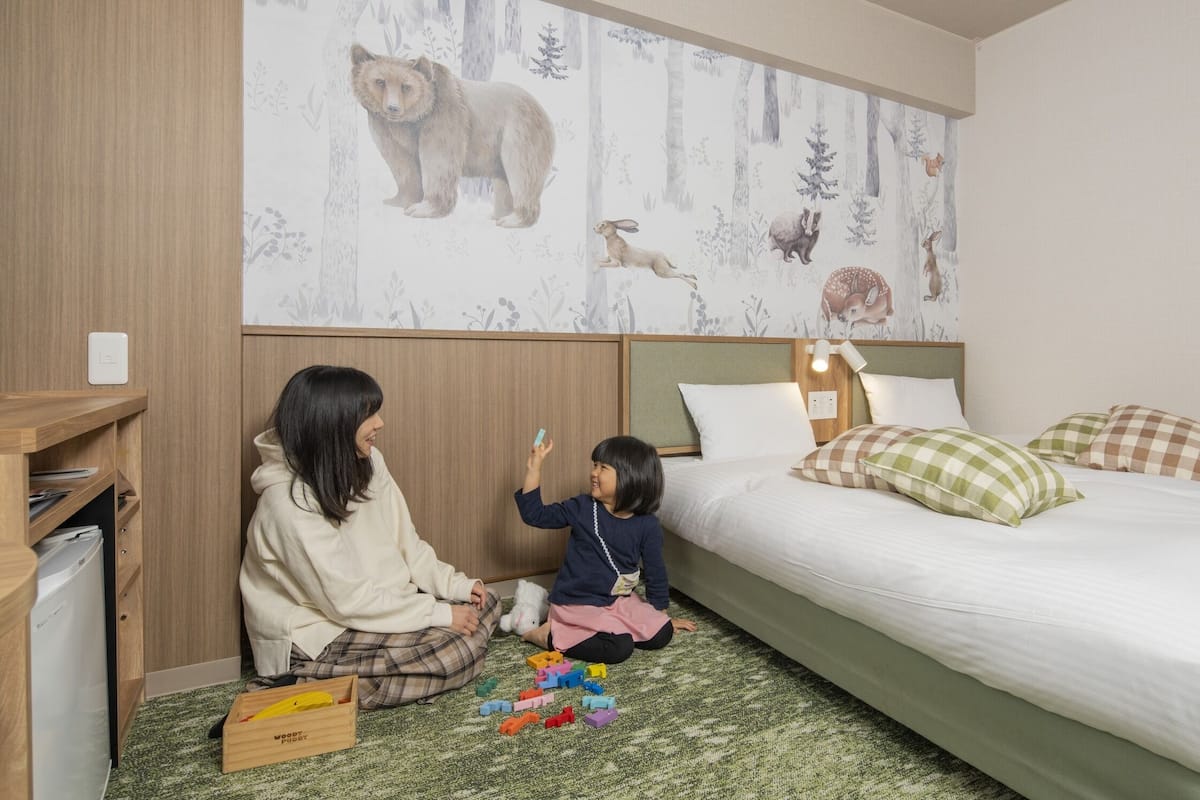 Une chambre à coucher comprenant un lit, une table de chevet, un mur recouvert d’un papier peint à motifs de forêt et une armoire en bois.
