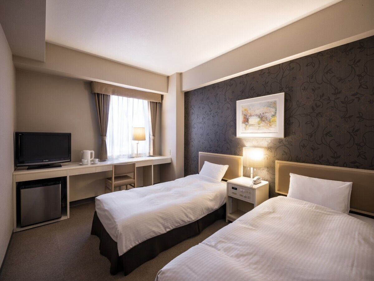 Une chambre d’hôtel avec deux lits, un bureau avec une télévision, une table de chevet avec une lampe et un petit réfrigérateur.