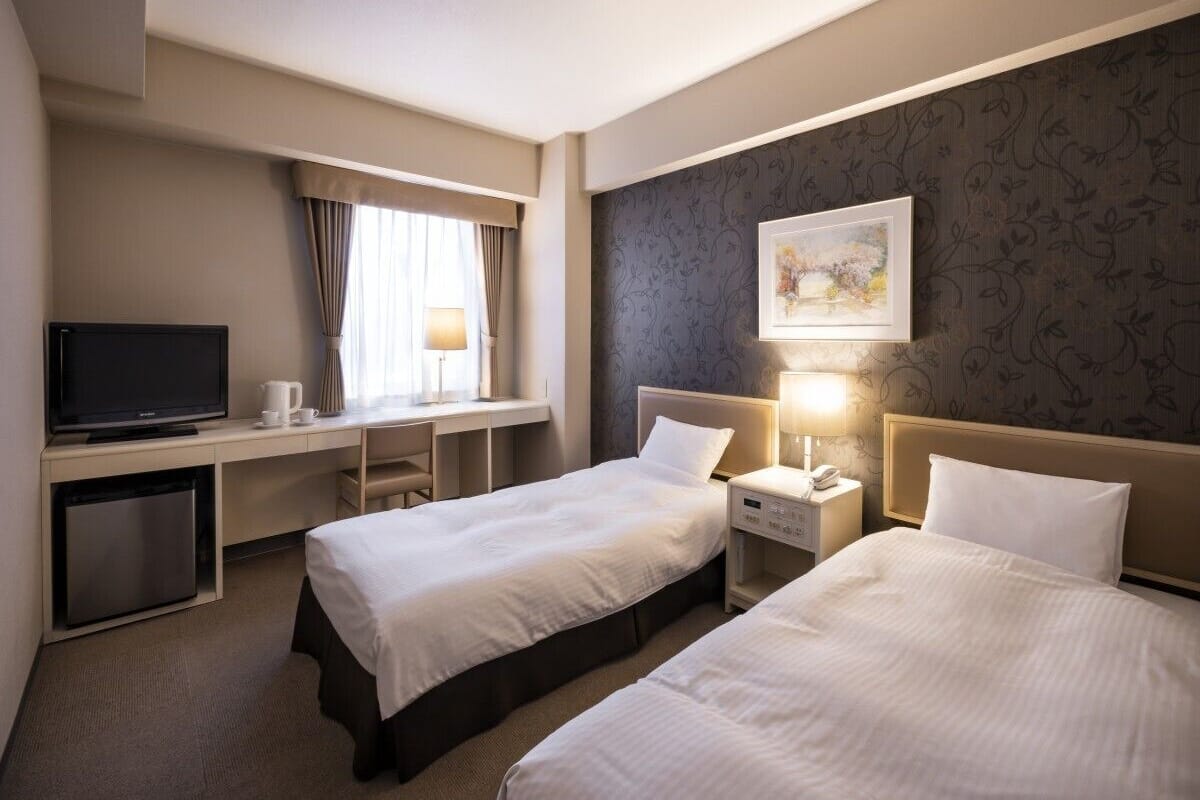 Une chambre d’hôtel avec deux lits, un bureau avec une télévision, une table de chevet avec une lampe et un petit réfrigérateur.