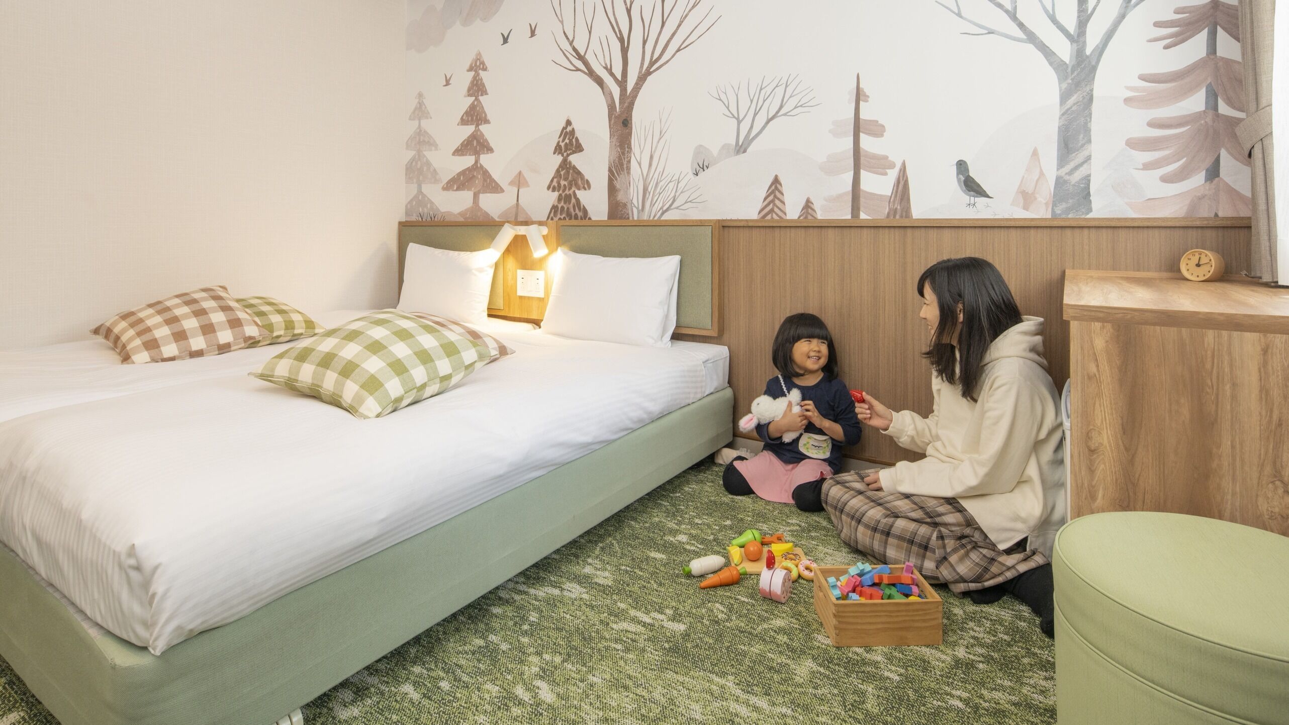 Un dormitorio con cama, almohadas, una mesita de noche, un cabecero de madera, un mural en la pared, un reloj y un baúl de juguetes.