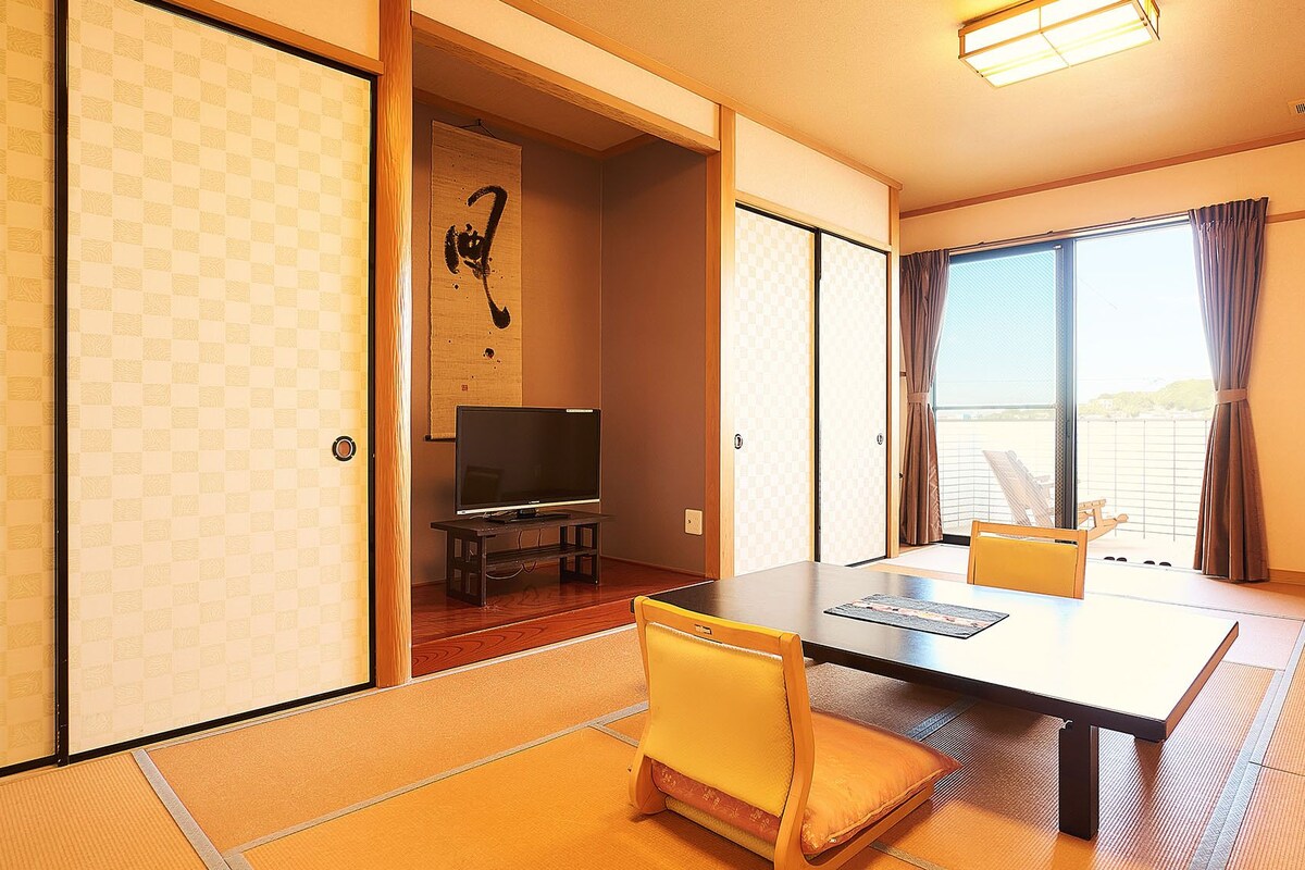 Une pièce de style japonais traditionnel, avec un sol recouvert de tatamis, un téléviseur posé sur un meuble bas et une porte coulissante donnant sur un balcon.