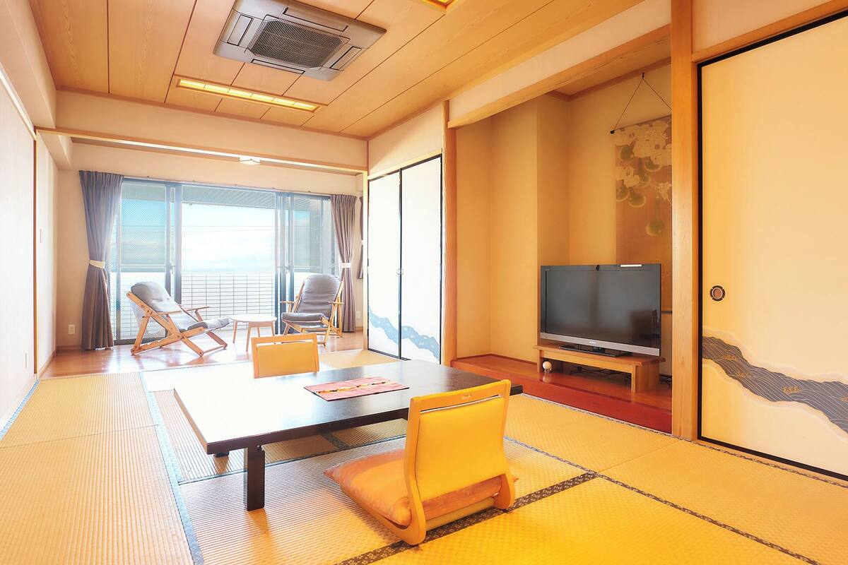 Ein Hotelzimmer mit Tatami-Boden, einer Couch, einem Fernseher und einer Schiebetür zu einem Balkon.