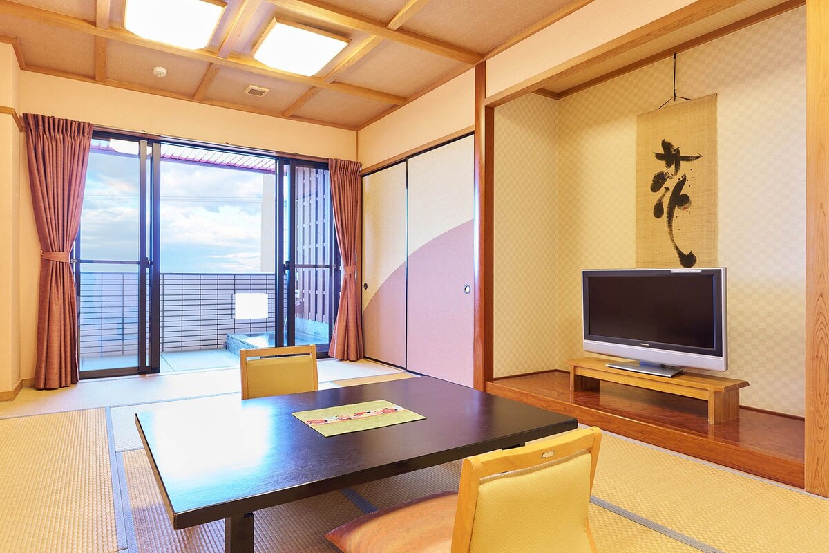A room with a large table, two chairs, a TV, and a sliding door to a balcony.